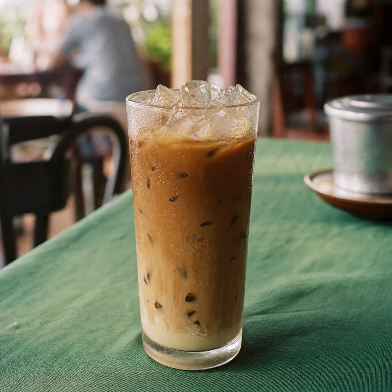 KI-generiert: Cà phê sữa đá im hohen Glas mit Eis und Kondenswasser auf einem grünen Tischtuch